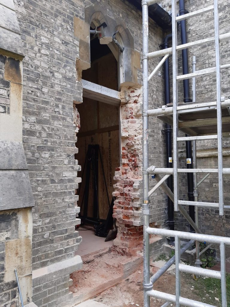 A photo looking into the church from the outside. It shows a very rough hole in the brickwork. the remains of the old windows can be seen at the top of the doorway and a lintel sits a small way down from the top of the gap. A scaffolding tower is in the right of the picture in front of the gap. 