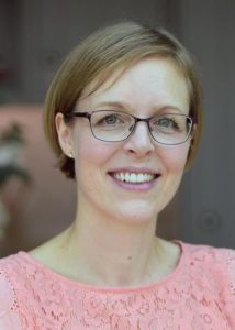 A woman with light brown hair wearing a light pink top and glasses.