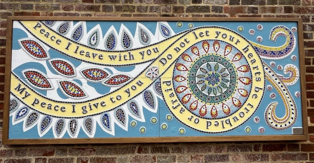 A rectangular mosaic art piece mounted on an old brick wall. The background is painted sky-blue and filled with stylized, teardrop-shaped leaf motifs in white, each inlaid with jewel-tone tiles of red, blue, green and yellow. A flowing yellow ribbon weaves across the panel, edged in black tiles, bearing the words in dark serif letters:
“Peace I leave with you
My peace I give to you
Do not let your hearts be troubled or afraid.”
To the right, a circular mandala of concentric rings of colorful tiles echoes the ribbon’s curve, and smaller dot motifs scatter around the edges.