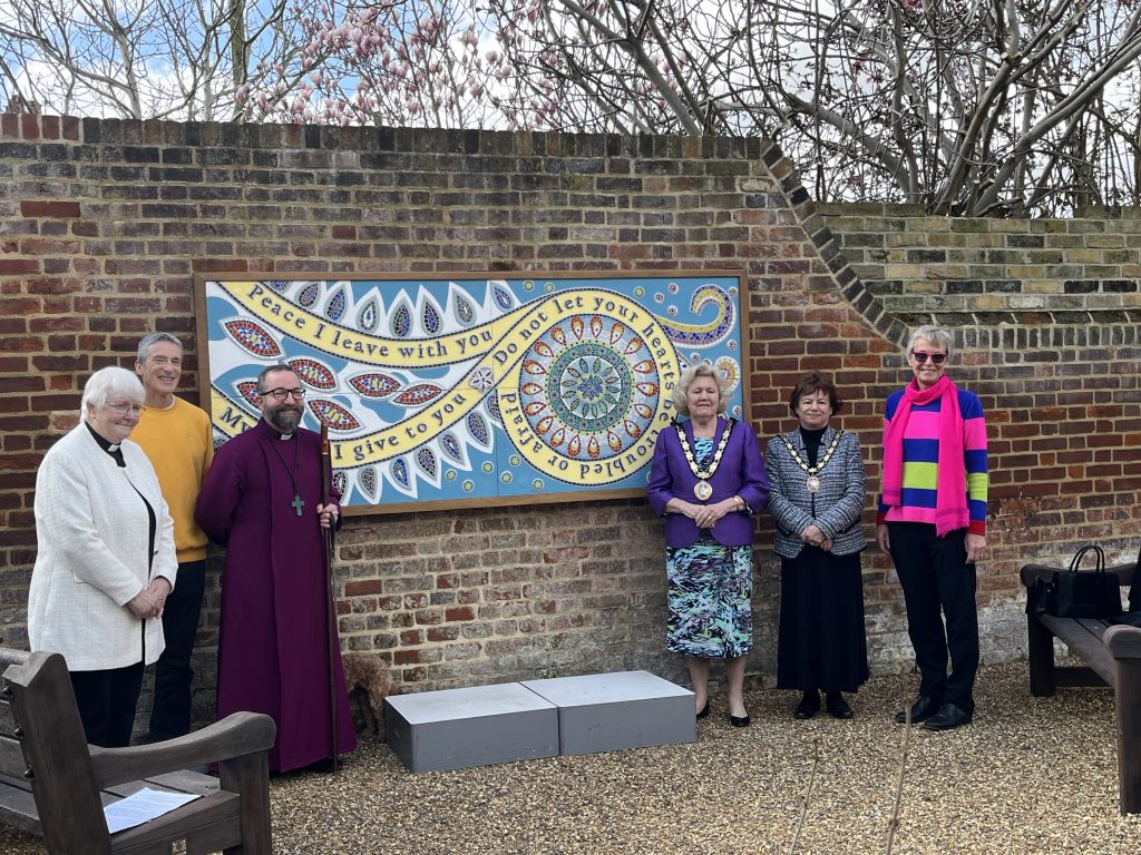 Six adults stand shoulder-to-shoulder in a gravel courtyard before the same mosaic panel mounted on a brick wall. From left to right: a woman in a light-colored clerical jacket; a man in a mustard sweater; a bishop in purple robes holding a staff; two women wearing formal dresses and mayoral chains; and a woman in a bright striped sweater and pink scarf. Behind them, a brick wall and leafless tree branches frame the scene.