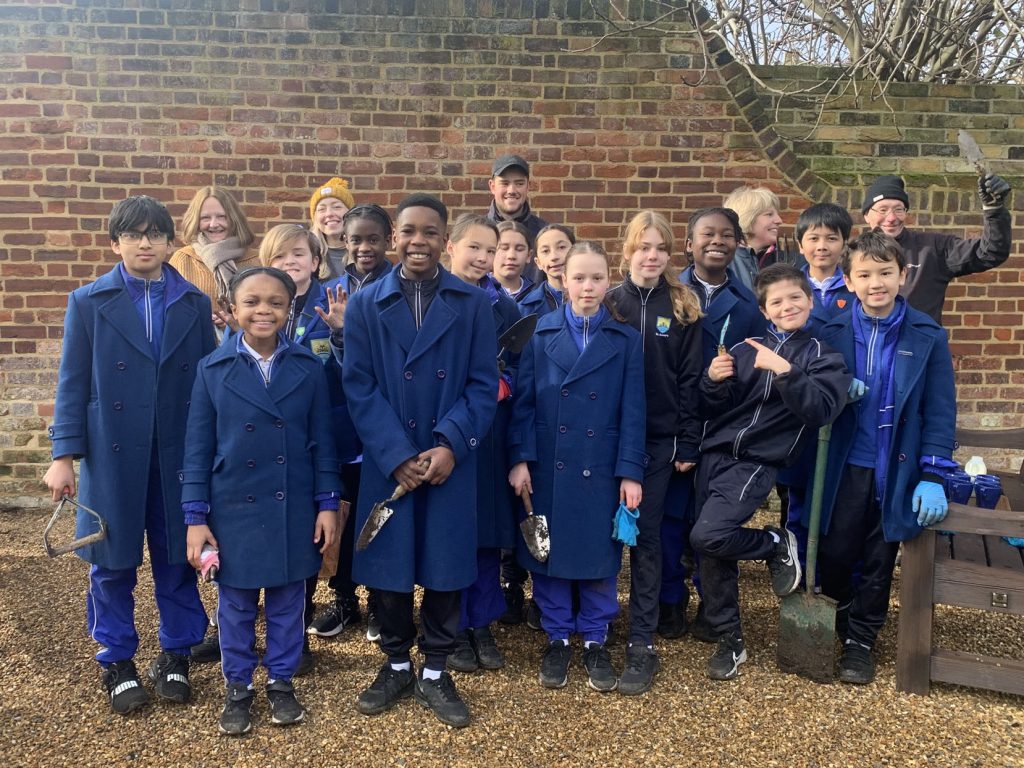 Group of smiling children and adults posing happily in front of a brick wall, wearing coats and gardening gloves, holding gardening tools.