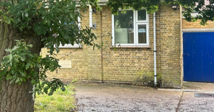An exterior view of the brick-built church hall, partly obscured by leafy branches of a tree. A window and a blue door are visible, set against a backdrop of greenery, conveying a peaceful and welcoming setting.