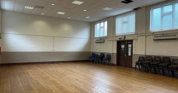 A large, open hall with polished wooden flooring and bright natural light entering through high windows. Chairs stacked neatly by a double door suggest flexible use, ideal for gatherings or activities.