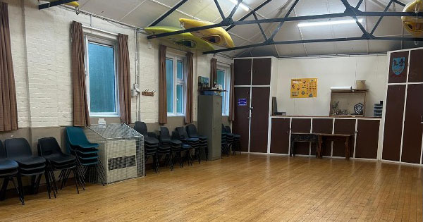 A multi-purpose community room with wooden floors, chairs neatly stacked along the walls, and two yellow kayaks stored overhead. Large windows and curtains line the side wall, giving the room a functional, welcoming appearance.
