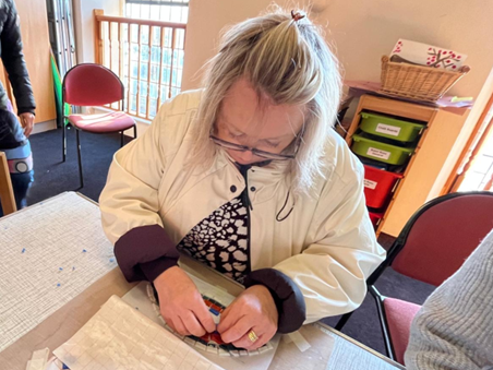 A woman sits ata a table placing mosaic tiles in a pattern. She is wearing glasses, a white coat and a black and white top 