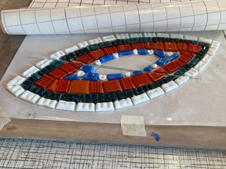 A section mosaic tiles sit on a slab on a table. The tiles are in the shape of an eye with white tiles on the outer edge, then a band of black tiles. a thicker band of red tiles and finally white and blue tiles towards the centre the middle is yet to be finished.
