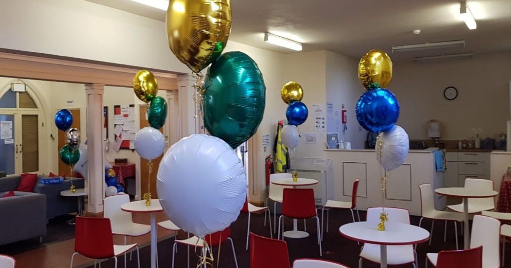 A warmly-lit lounge area decorated for a celebration, featuring round tables and modern chairs. Bright helium balloons in gold, green, blue, and white float elegantly above the tables, creating a festive atmosphere.