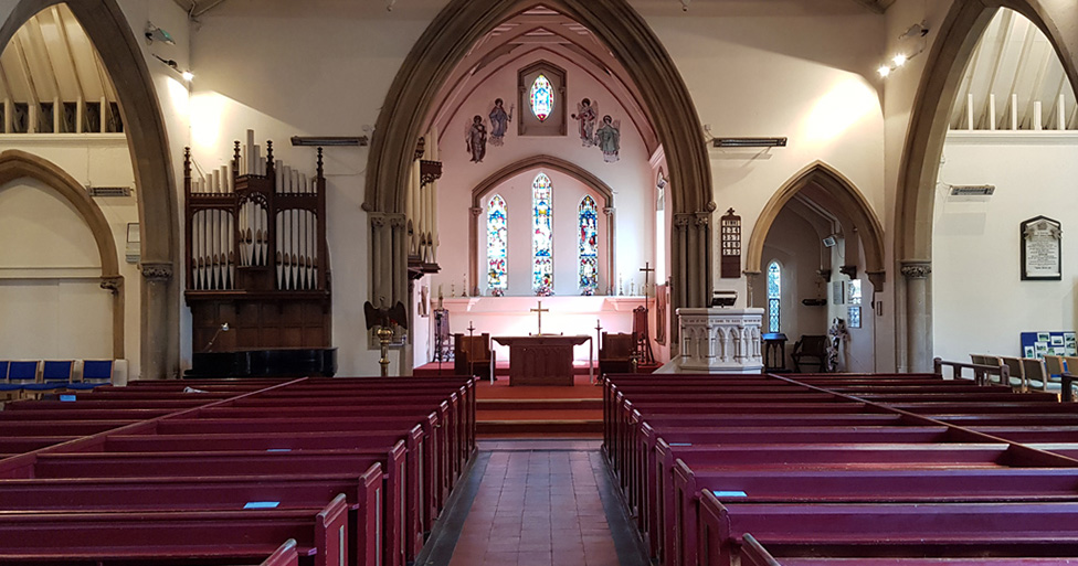 An interior view of the church, with rows of red pews leading to an altar beneath arched ceilings. Stained glass windows and decorative artwork adorn the walls, with a traditional church organ visible to the left.