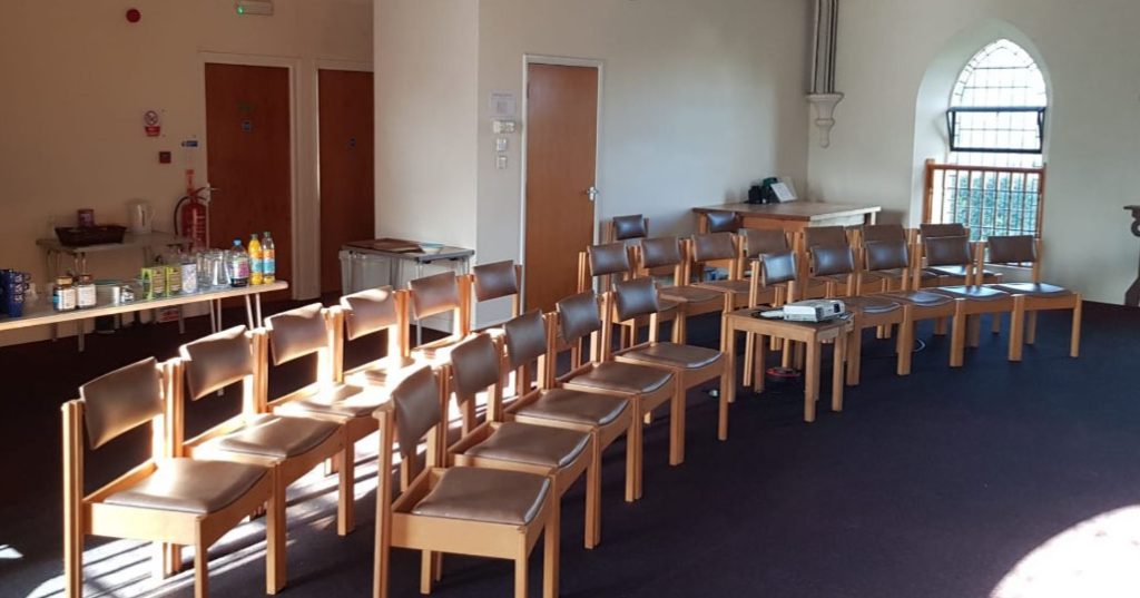 A sunlit meeting room with rows of chairs arranged in a gentle curve. A small projector is placed on a table among the chairs. Refreshments and cups are laid out on a side table, suggesting preparation for a meeting or workshop.