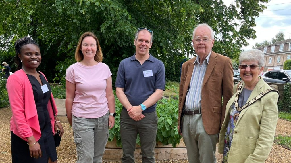 Five adults pose and smile for a photo beside a raised garden bed. They are wearing name badges. They include the Mp for Chelmsford Marie Goldman and Mayor of Chelmsford Councillor Jannetta Sosin. Behind them is a tree-lined gravel path and residential buildings in the distance.