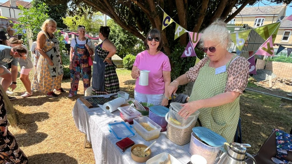 Two women stand behind a table covered in containers of food and drink, offering refreshments at the green fair. Other attendees chat nearby, under bunting. The setting is sunny with dappled tree shade.