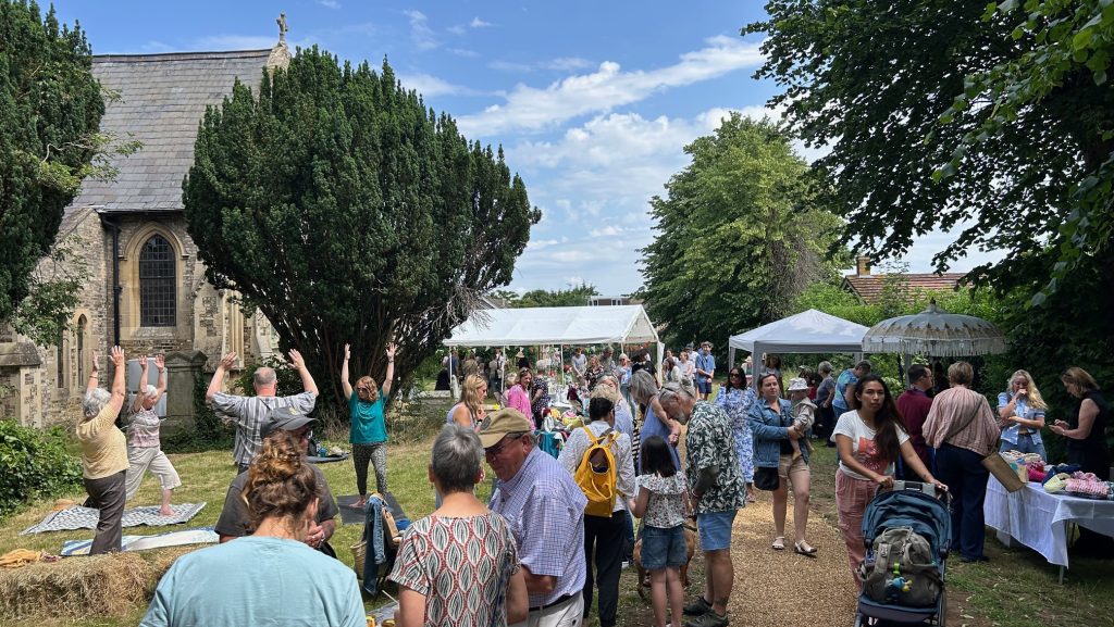 A lively crowd enjoys a summer fair in the grounds of a church, with stalls, yoga participants, and people browsing items under white gazebos. Trees and a blue sky frame the cheerful, bustling atmosphere.