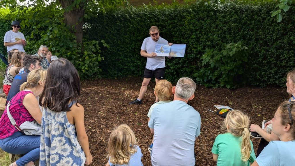 A man in sunglasses reads aloud from a picture book to a group of children and adults seated outdoors on woodchip ground under trees, with dense green hedging in the background. The scene has a casual, community feel.