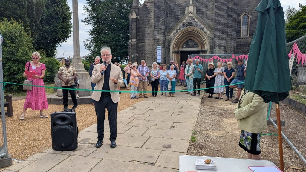 Archdeacon Jonathan Croucher speaks into a microphone at a ribbon-cutting ceremony outside St John's church. A crowd of people stands behind him, and pink bunting decorates the scene. A woman in a pink dress stands to the side reading from notes.