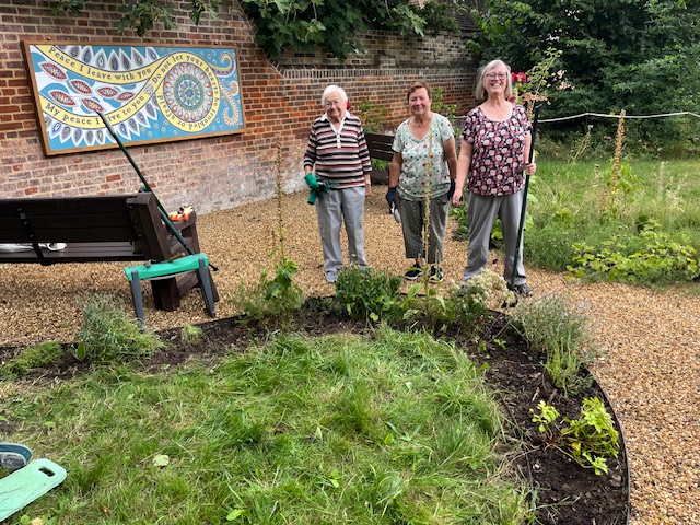 Elizabeth, Christine and Jacky from our congregation in the Green Space, holding gardening tools and smiling, with a bench, a decorative mural on a brick wall, and newly planted greenery in front of them.